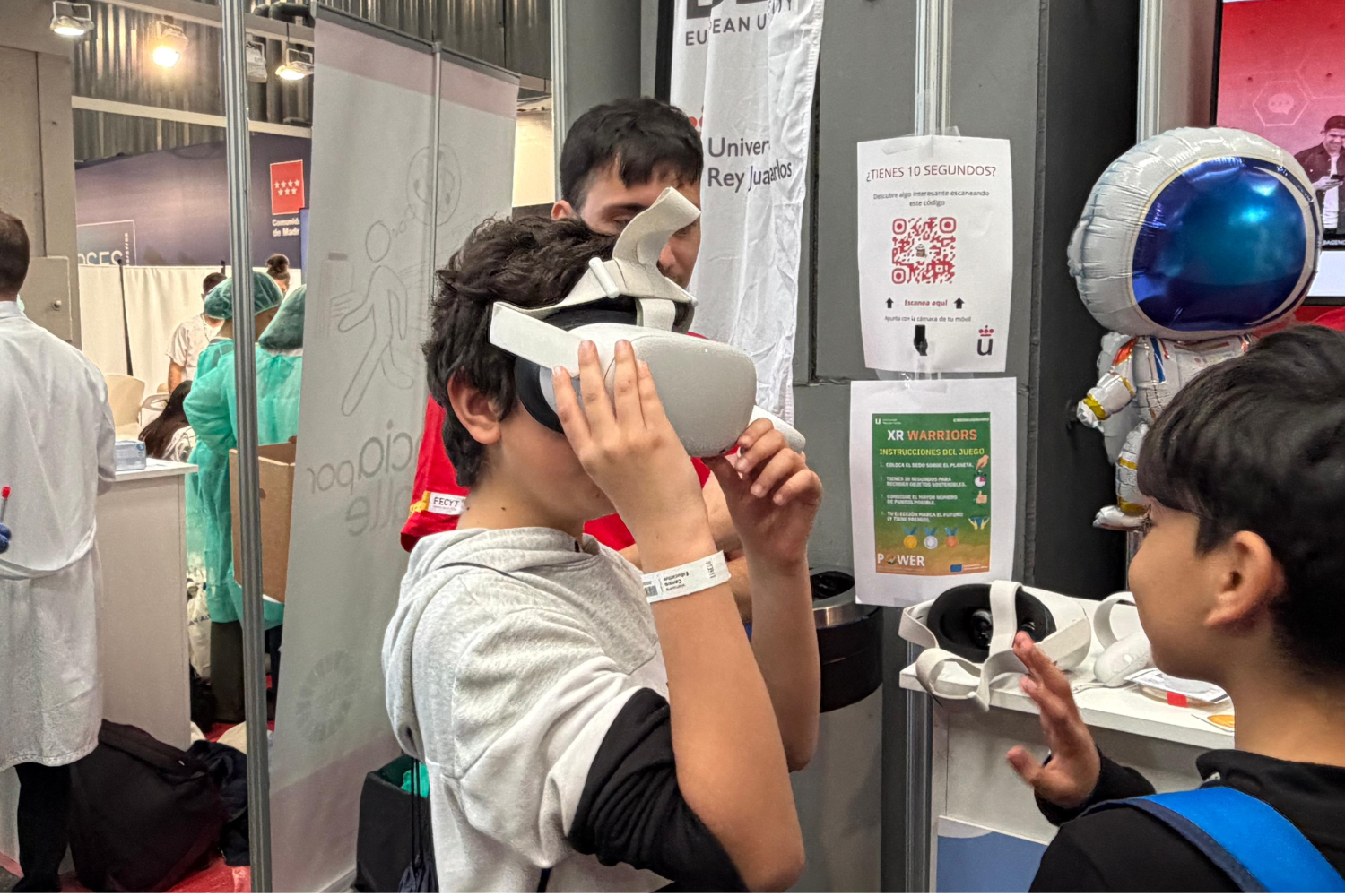Un grupo de niños participa en una actividad en un stand de feria científica. En primer plano, un niño se coloca unas gafas de realidad virtual mientras otro observa y le habla. Detrás, se ve el puesto con carteles informativos, varios dispositivos de VR sobre una mesa y un muñeco de astronauta decorativo. Al fondo, otras personas con batas participan en actividades relacionadas con ciencia o salud.