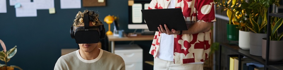 Young adult Caucasian man wearing virtual reality headset sitting in modern office while middle aged man holding laptop standing nearby conducting technology demonstration 