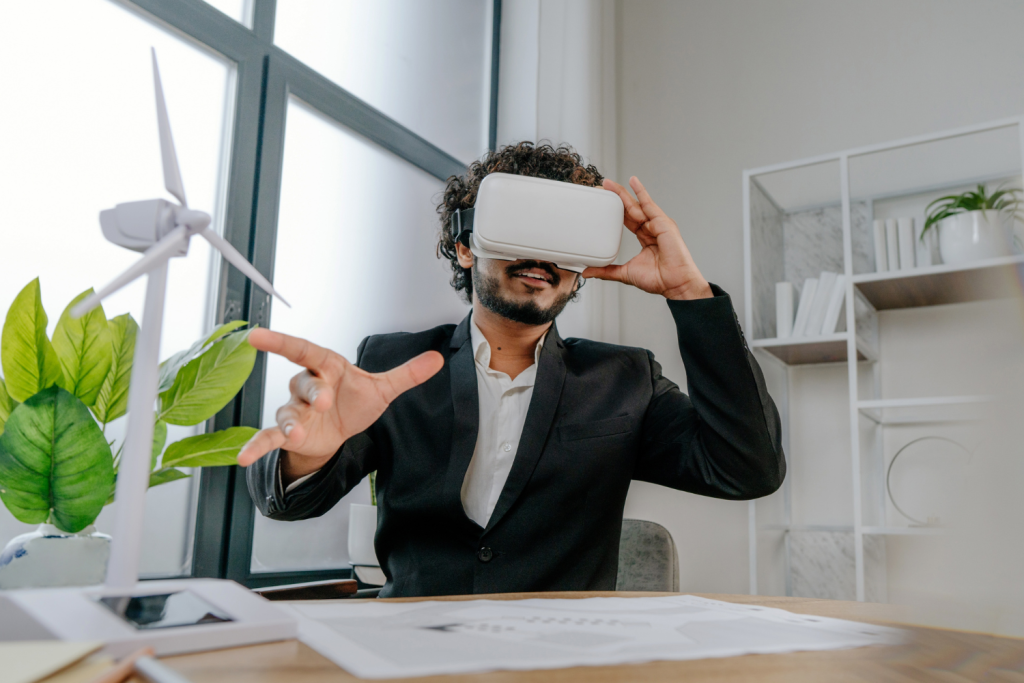 Hombre con gafas de realidad virtual en un escritorio moderno, interactuando con un entorno digital; a su lado, una maqueta de aerogenerador y una planta sugieren sostenibilidad.