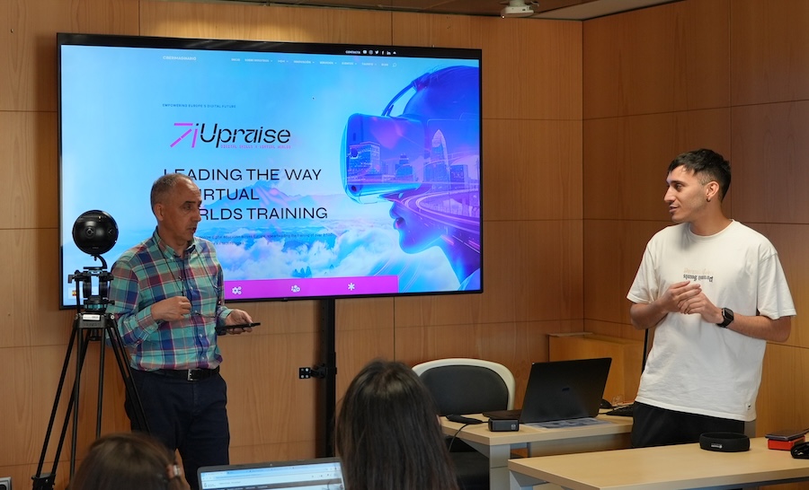 Two instructors lead a classroom session on immersive technologies and virtual worlds training in front of a large screen displaying the UPRAISE platform. Students work on laptops while a 360-degree camera mounted on a tripod suggests a hands-on workshop related to XR, immersive journalism, or digital storytelling.