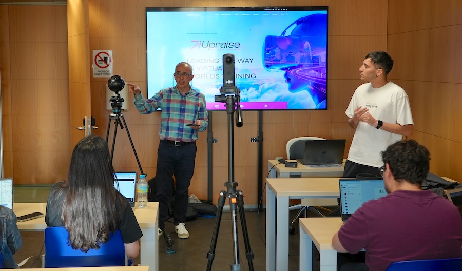 Grupo de personas en una sesión formativa sobre tecnologías inmersivas y realidad virtual en un aula. Dos ponentes presentan frente a una pantalla con el logotipo de “iUprize”, mientras varios asistentes trabajan con ordenadores portátiles. En primer plano hay cámaras y equipos de grabación sobre trípodes, sugiriendo una demostración o actividad relacionada con XR y producción audiovisual.